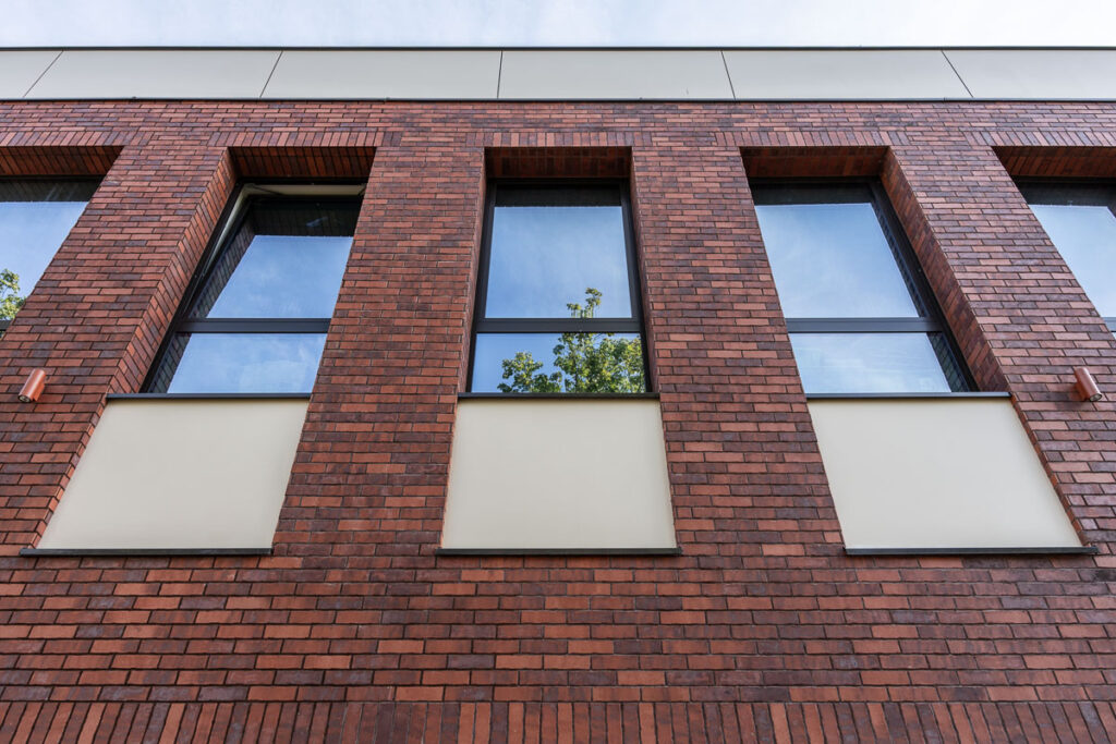 Ceglana elewacja nowoczesnego budynku z dużymi oknami odbijającymi niebo i drzewa - Fotografia Architektury Wrocław