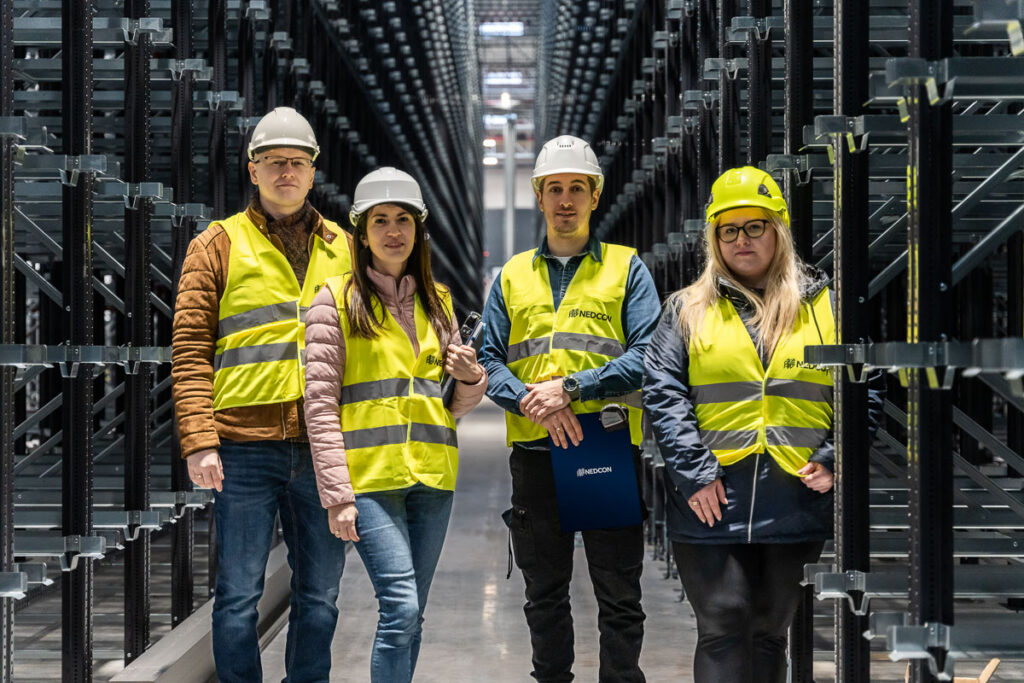 Grupa pracowników w żółtych kamizelkach i białych kaskach z regałami pojemnikowymi w tle - Fotografia Biznesowa Wrocław