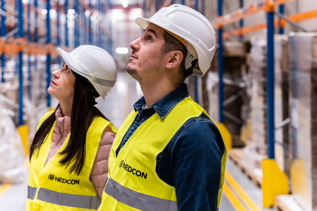 mężczyzna i kobieta w żółtych kamizelkach i białych kaskach pomiędzy regałami magazynowymi - Fotografia Biznesowa Wrocław